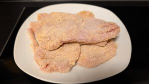 Breaded raw cutlets in pale orange crumbs stacked on a white plate before frying