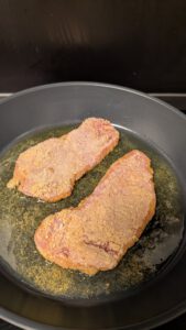 Two breaded cutlets frying in a shallow layer of fat in a large dark pan, the crumb coating still pale at the edges