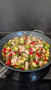 Large pan on the stove with chicken pieces, broccoli, red pepper, zucchini and sauteed mushrooms mixed together