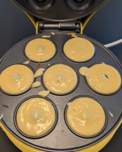 Donut batter filled into the seven wells of an open yellow electric donut maker, with a few drips on the plate