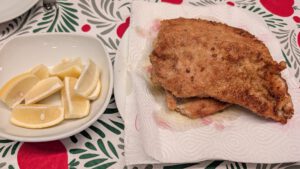 Two fried golden-brown schnitzels on paper towel next to a white bowl of lemon wedges, on a red and green patterned tablecloth