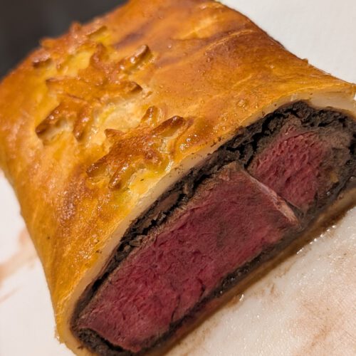 Cross-section of a baked Beef Wellington with pink beef, a dark duxelles layer and golden pastry with a snowflake decoration on a white board with a green rim