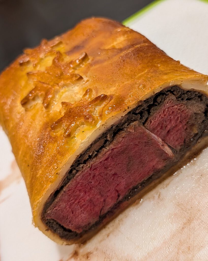 Cross-section of a baked Beef Wellington with pink beef, a dark duxelles layer and golden pastry with a snowflake decoration on a white board with a green rim