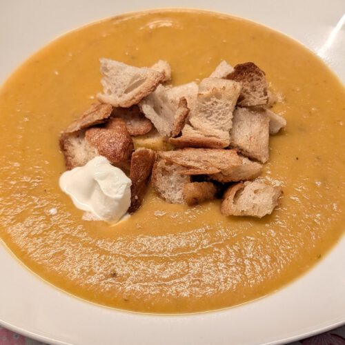 Creamy orange carrot potato soup in a white square bowl topped with golden croutons and a dollop of sour cream on a heart-patterned tablecloth