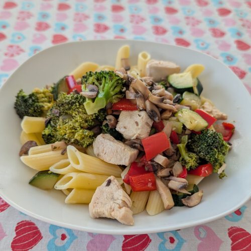White plate of penne with chicken pieces, broccoli, red pepper, zucchini and sliced mushrooms on a heart-patterned tablecloth