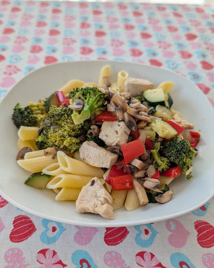 White plate of penne with chicken pieces, broccoli, red pepper, zucchini and sliced mushrooms on a heart-patterned tablecloth