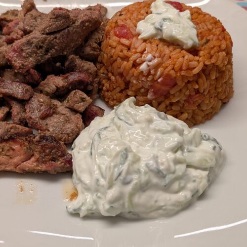 White square plate with sliced seared gyros meat, a dome of tomato rice topped with tzatziki, and a mound of tzatziki with cucumber