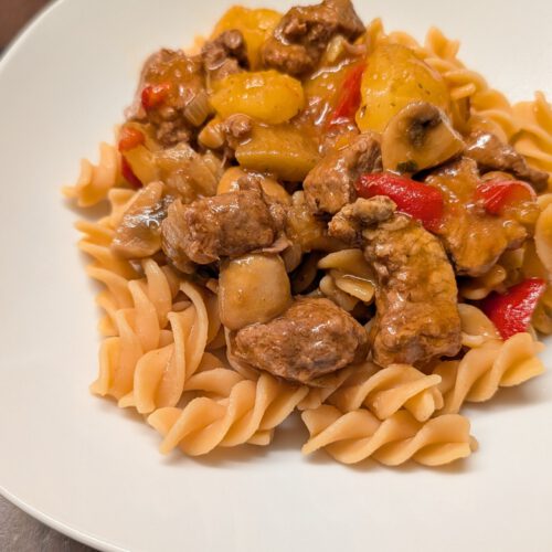Beef goulash with potato chunks, mushrooms and red bell pepper pieces on spiral pasta on a white plate on a dark wooden table