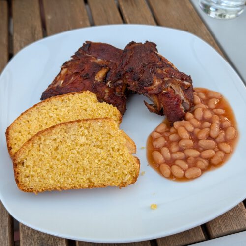 White plate with glazed BBQ spare ribs, two slices of golden cornbread and baked beans in tomato sauce on a wooden outdoor table