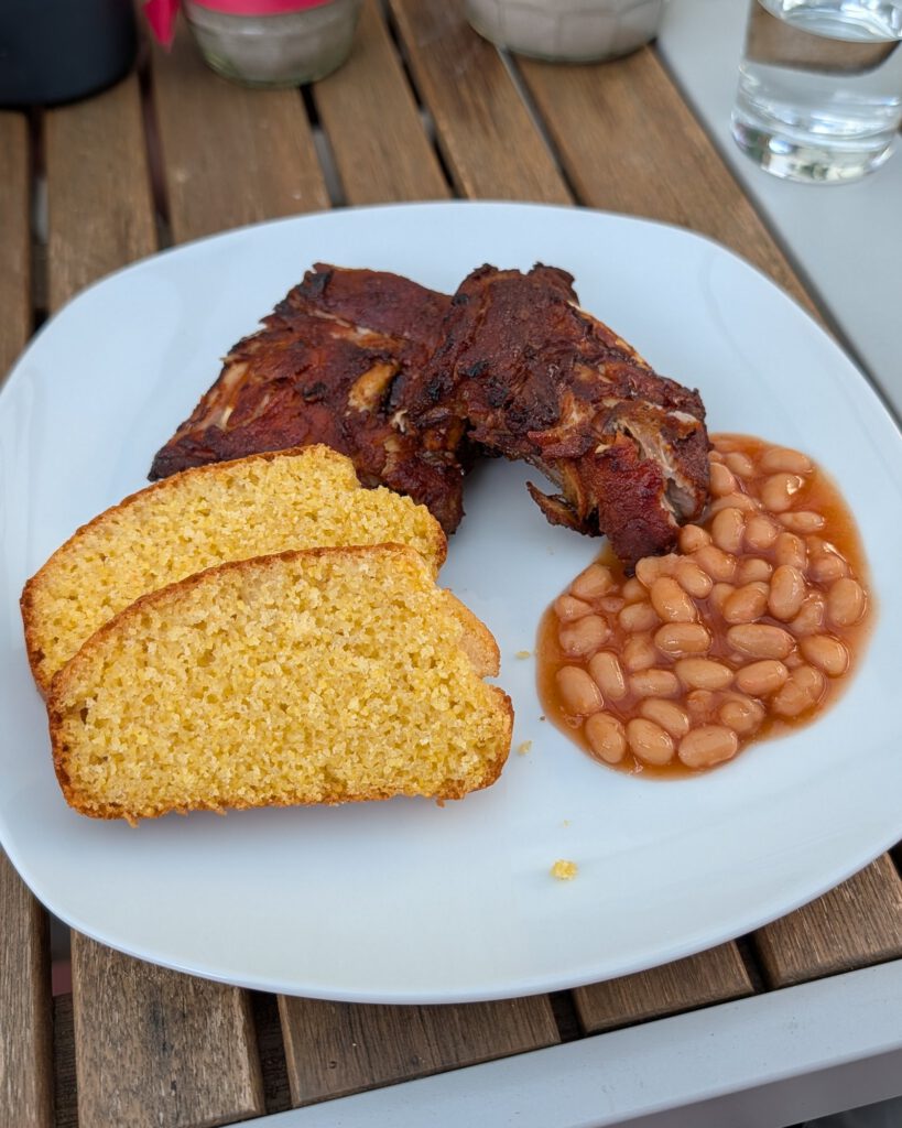 White plate with glazed BBQ spare ribs, two slices of golden cornbread and baked beans in tomato sauce on a wooden outdoor table