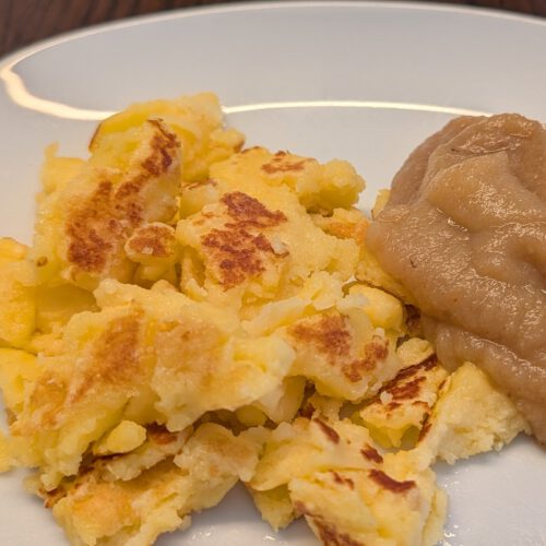 Golden shredded Kaiserschmarrn pieces and a mound of apple sauce on a white plate on a dark wooden table
