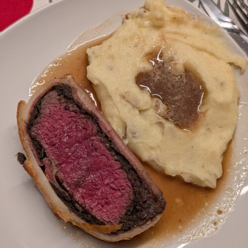Creamy mashed potatoes topped with brown sauce on a plate next to a slice of Beef Wellington