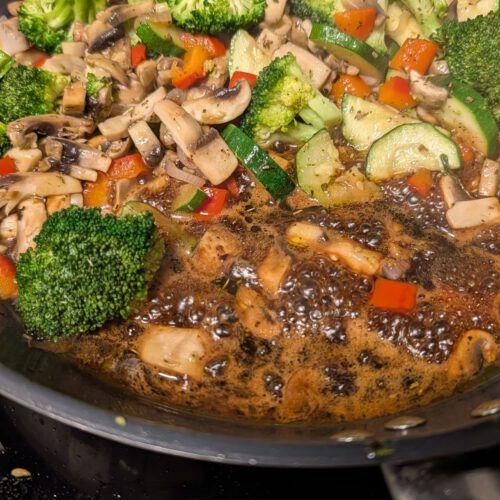 Frying pan full of broccoli florets, sliced mushrooms, zucchini slices and red bell pepper pieces with dark balsamic syrup bubbling at the bottom