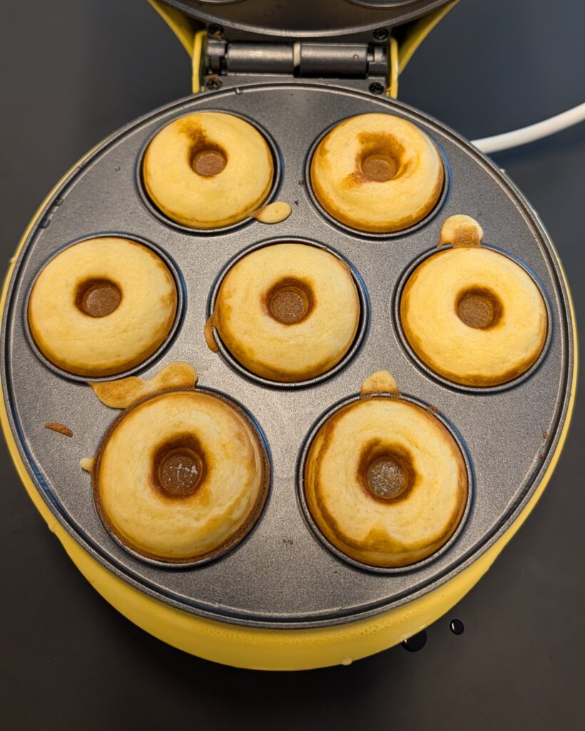 Seven freshly baked golden mini donuts in the open wells of a yellow electric donut maker, seen from above