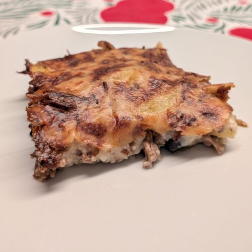 Piece of baked moussaka on a white plate with a browned top with dark speckles; ground beef and pale creamy layers are visible at the cut side