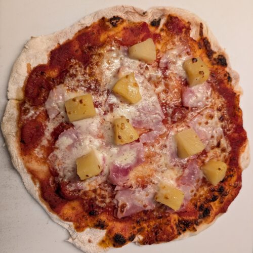 Baked pizza with ham, melted cheese, tomato sauce and pineapple chunks on a white baking surface
