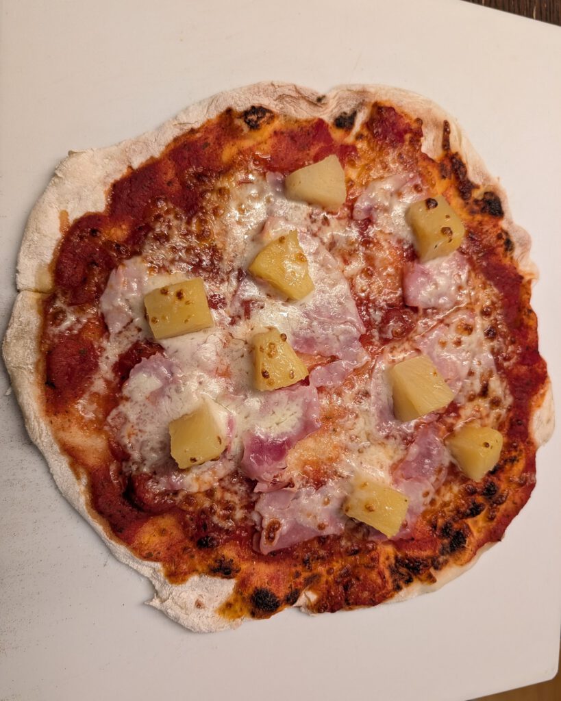 Baked pizza with ham, melted cheese, tomato sauce and pineapple chunks on a white baking surface