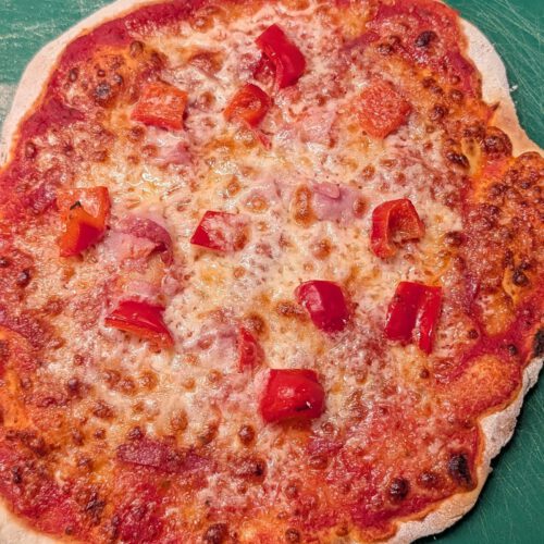 Baked gluten-free pizza with tomato sauce, melted mozzarella, pieces of ham and diced red bell pepper on a green board