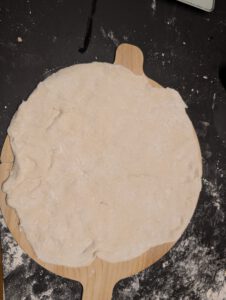 Pizza dough pressed flat by hand on a wooden pizza peel with flour on a dark worktop