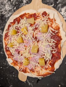 Raw topped pizza on a pizza peel with tomato sauce, ham, pineapple chunks and shredded cheese