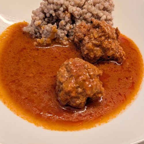 White square plate with two meatballs covered in tomato sauce, a pile of cooked buckwheat groats and a pool of orange-red tomato sauce on a polka-dot tablecloth