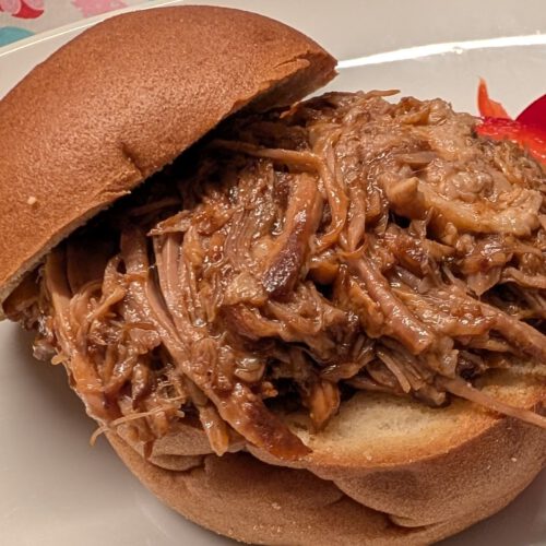 Pulled pork on a gluten-free bun with red bell pepper strips on a white plate, heart-patterned tablecloth behind