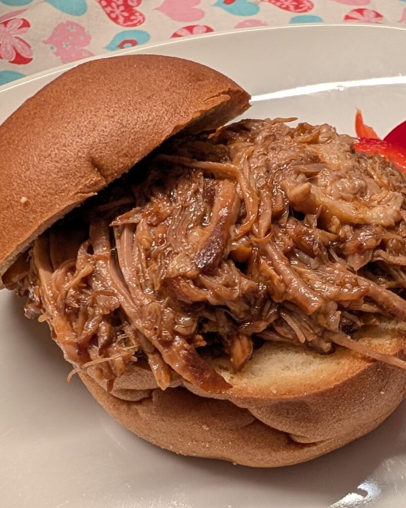 Pulled pork on a gluten-free bun with red bell pepper strips on a white plate, heart-patterned tablecloth behind