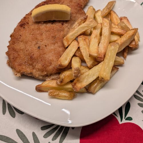 Breaded schnitzel with a golden-brown crumb crust on a white plate with a lemon wedge on top and a pile of golden fries beside it, on a red, green and white patterned tablecloth