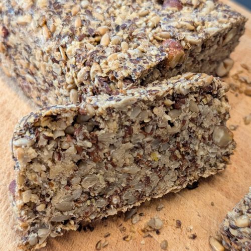 Seed loaf with one thick slice cut off and laid in front, showing the crumb of sunflower seeds, flaxseed and hazelnuts, on a wooden board