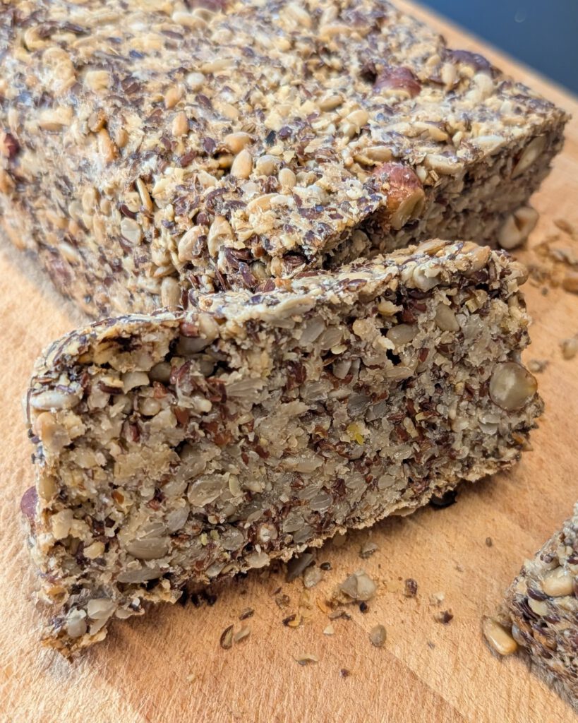 Seed loaf with one thick slice cut off and laid in front, showing the crumb of sunflower seeds, flaxseed and hazelnuts, on a wooden board
