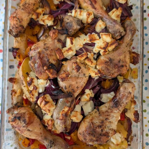 Baked chicken drumsticks in a rectangular glass dish with potato wedges, red onion and bell pepper, topped with browned feta crumbles