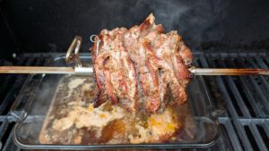 Marinated pork slices packed on a rotisserie spit inside a gas grill with a glass dish of drippings underneath