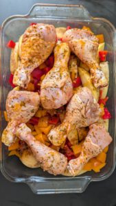 Marinated raw chicken drumsticks lying on potato slices and diced red and orange bell pepper in a rectangular glass baking dish
