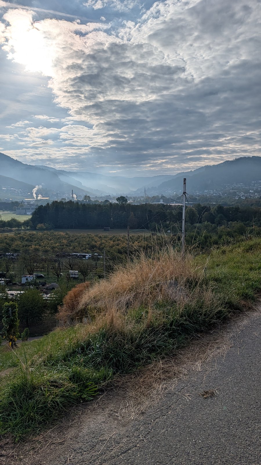 Gravel path with dry grass and a wooden post on a hillside, a hazy valley with a small town and forested hills under a cloudy sky