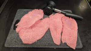 Four raw pink cutlets pounded flat on a dark gray cutting board, with a black meat mallet lying behind them