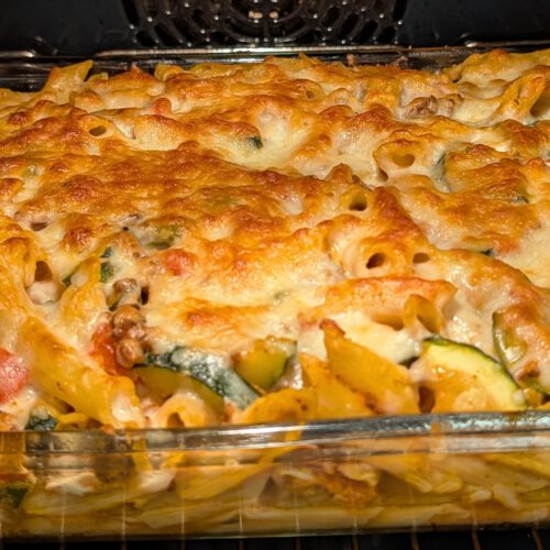 Baked penne casserole in a rectangular glass dish inside the oven, with a golden-brown bubbling cheese crust over penne and zucchini