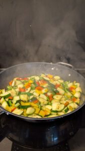 Diced zucchini and red, yellow and green bell pepper searing in a large steaming pan on a cooktop