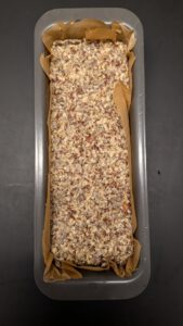 Seed bread dough pressed flat into a parchment-lined loaf pan, seen from above, ready for the overnight rest in the refrigerator