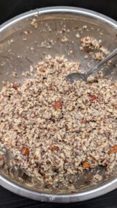 Thick seed bread batter with a spoon in a steel bowl, oats, seeds, nut pieces and grey chia seeds visible