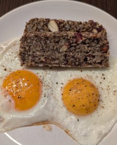 Two stacked slices of seed bread with nuts above two sunny-side-up eggs with pepper on a white plate