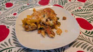 One serving of penne casserole on a white plate with browned cheese crust, melted cheese strands, ground beef sauce and zucchini, on a patterned tablecloth with red apples