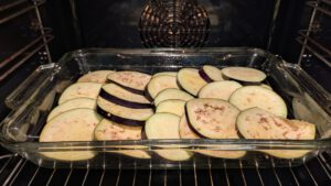 Raw eggplant slices with purple skin stacked in an overlapping layer in a rectangular glass baking dish inside the oven; no potato slices are visible