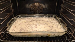 Unbaked moussaka in a rectangular glass baking dish in the oven, covered with a pale creamy sauce with crumbs of ground beef showing through